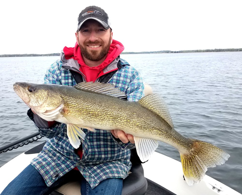 A smiling man wearing a plaid jacket and red hoodie sits on a boat, holding a large walleye fish during the fishing opener, with a lake and cloudy sky in the background.
