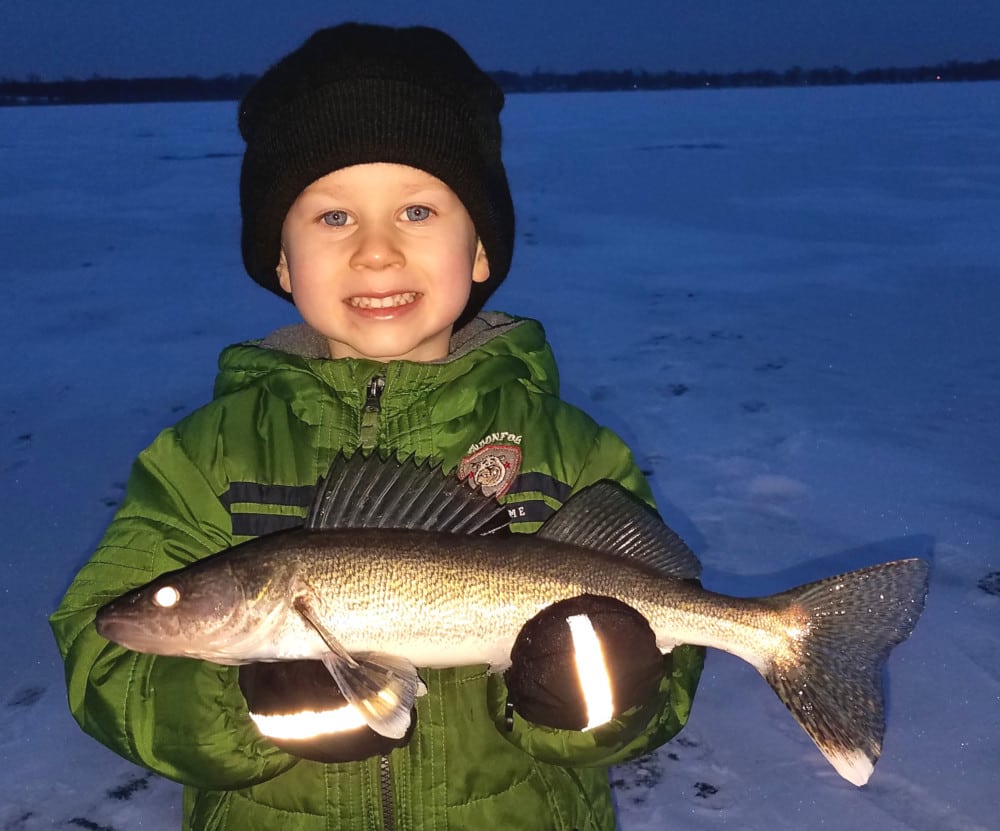 A young child in a green jacket and black hat smiles while holding a large walleye with both hands, standing on a snowy, frozen lake near Alexandria at dusk.