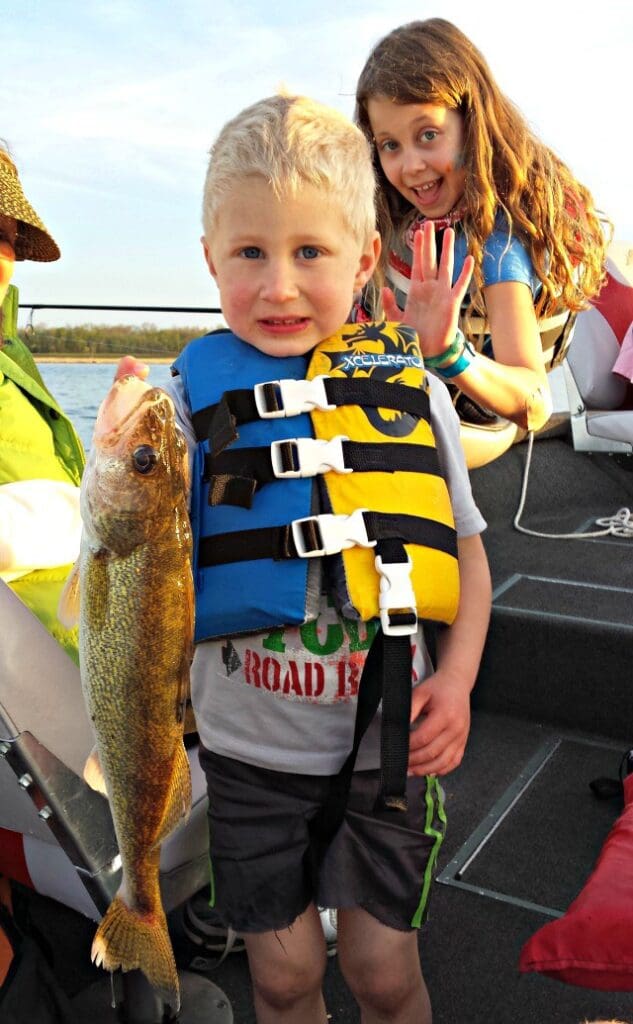 A young boy wearing a blue and yellow life jacket proudly holds a large fish on a boat, while a smiling girl in the background flashes a "four" hand sign and poses cheerfully—capturing the spirit of an early summer fishing report for Alexandria.
