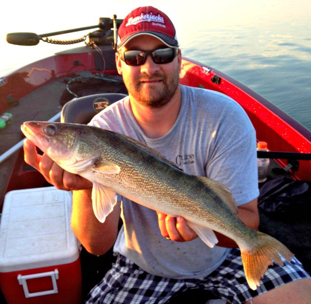A man wearing sunglasses, a red cap, and a gray t-shirt sits in a red boat holding a large fish. Smiling for the Alexandria fishing report for July, he poses with water in the background and a white cooler beside him.