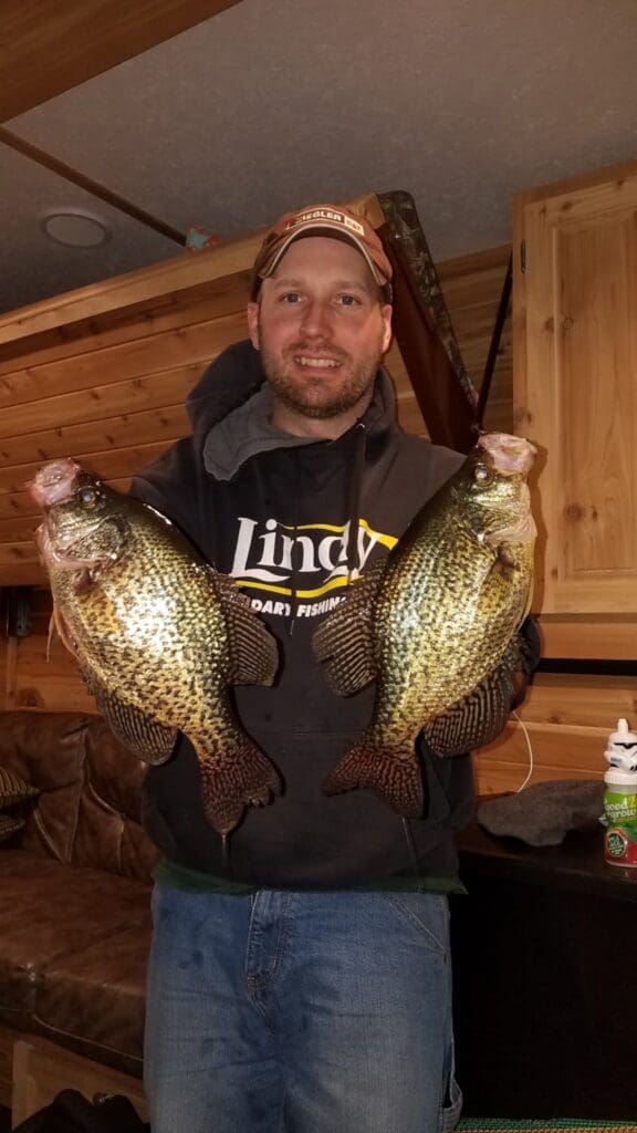 A man in a hoodie and cap smiles while holding two large crappie fish indoors, with wooden walls and fishing gear in the background—showcasing a successful fishing day in Alexandria.