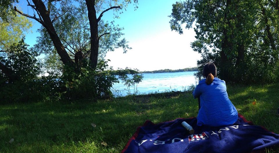 A person in a blue shirt sits on a dark blanket, enjoying a picnic in a park near a calm lake, surrounded by trees and grass with hints of fall color, gazing at the water under a clear, sunny sky.
