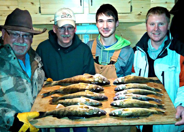 Four men indoors smile at the camera while holding a wooden board with ten freshly caught fish arranged in two rows—a great catch featured in this January fishing report. The background showcases wooden cabinetry, highlighting an Alexandria fishing success.