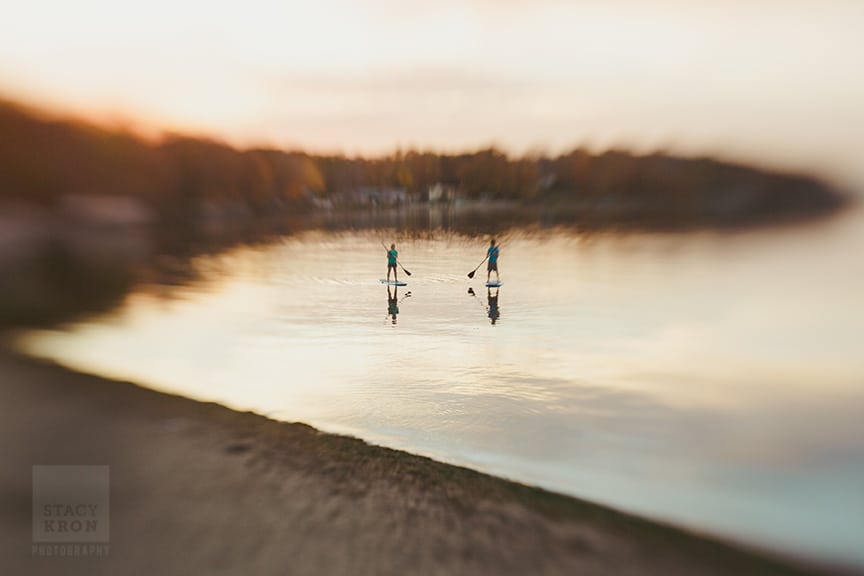Two people stand on paddleboards in calm water at sunset, each holding a paddle, as soft focus photography blurs the surroundings and reflects the warm sky and shoreline in the background.