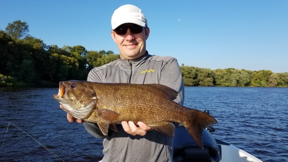 A man wearing sunglasses and a white cap holds a large brown fish while standing on a boat in the lake, enjoying the aggressive fall bite in the Alexandria Lakes area, with trees and a clear blue sky in the background.