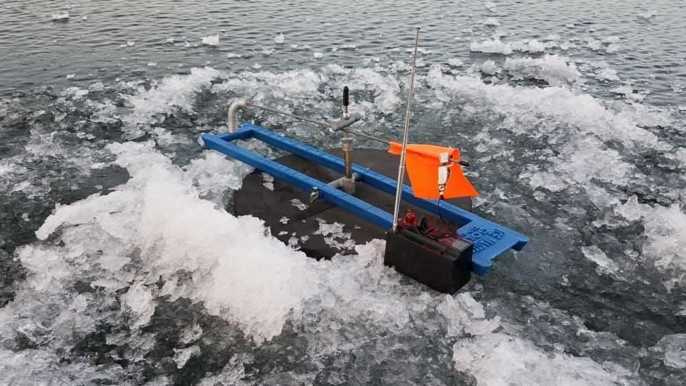 A small blue and black robotic device with an orange flag floats among chunks of ice on the surface of a body of water in the Alexandria area.