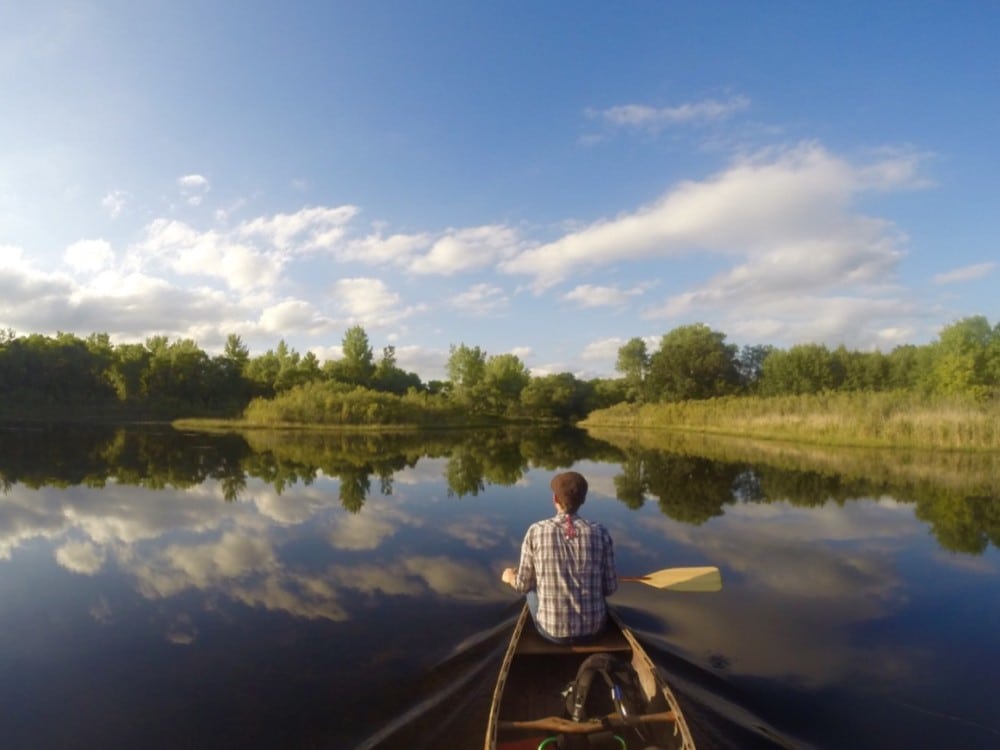 A person in a plaid shirt paddles a canoe on a calm lake in the Alexandria Lakes Area, surrounded by trees and greenery, with blue sky and clouds mirrored on the water—one of the 20 must-do summer activities in this scenic region.