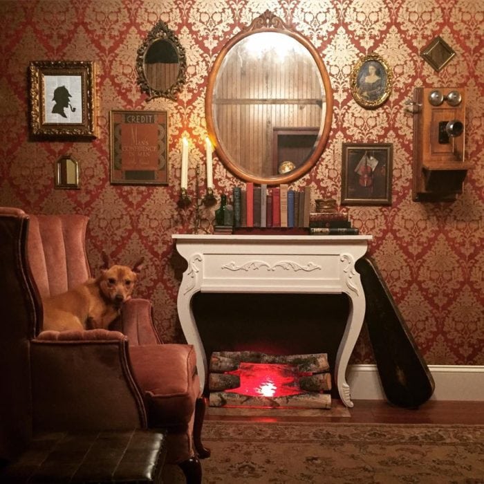 A cozy vintage Escape Room Alexandria with ornate red wallpaper, a faux fireplace, antique decor, framed pictures, books, and a brown dog sitting on a velvet armchair. Logs with red lights imitate a fire in the fireplace.