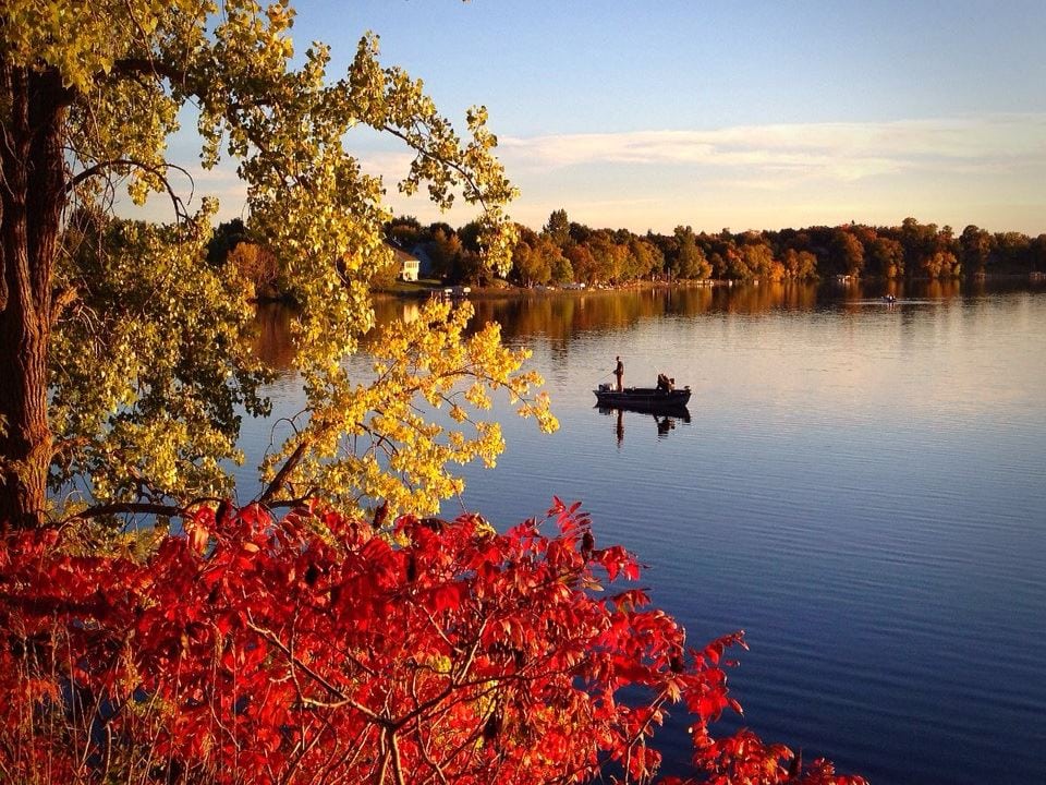 A calm lake reflects autumn trees with red, yellow, and green leaves. Two people fish in a small boat under a clear blue sky—just one of the 15 ways to explore Alexandria MN in October.