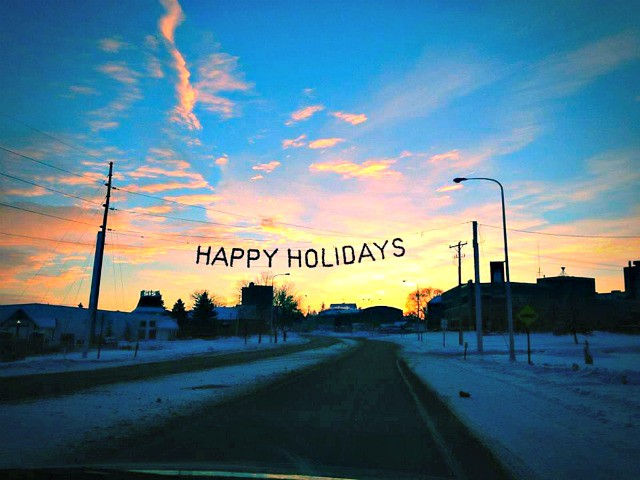 A snowy street in Alexandria at sunset, with buildings on either side and a "HAPPY HOLIDAYS" banner stretched across the road. The colorful sky and festive scene invite all to celebrate the season amid blue, orange, and pink clouds.