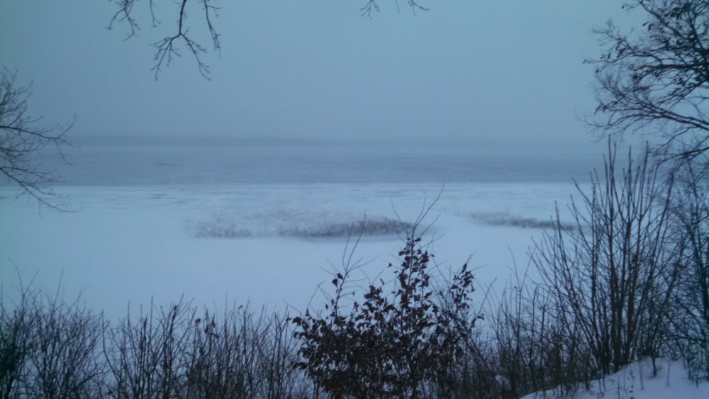 A frozen lake, ice beginning to form on area lakes, lies covered in snow under a cloudy, gray sky, with bare trees and shrubs in the foreground and branches framing the scene.