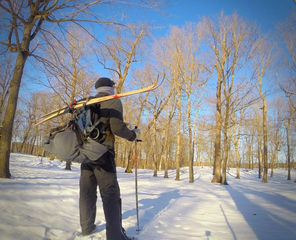 A person in winter clothing stands in a snowy forest near Alexandria, holding ski poles and carrying skis and a large backpack. Bare trees surround them under a clear blue sky.