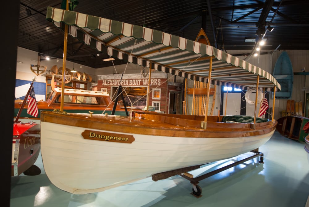 A vintage wooden boat named "Dungeness" with a striped canopy is displayed indoors at the Legacy of the Lakes Museum, with an American flag on the stern and an "Alexandria Boat Works" sign in the background.