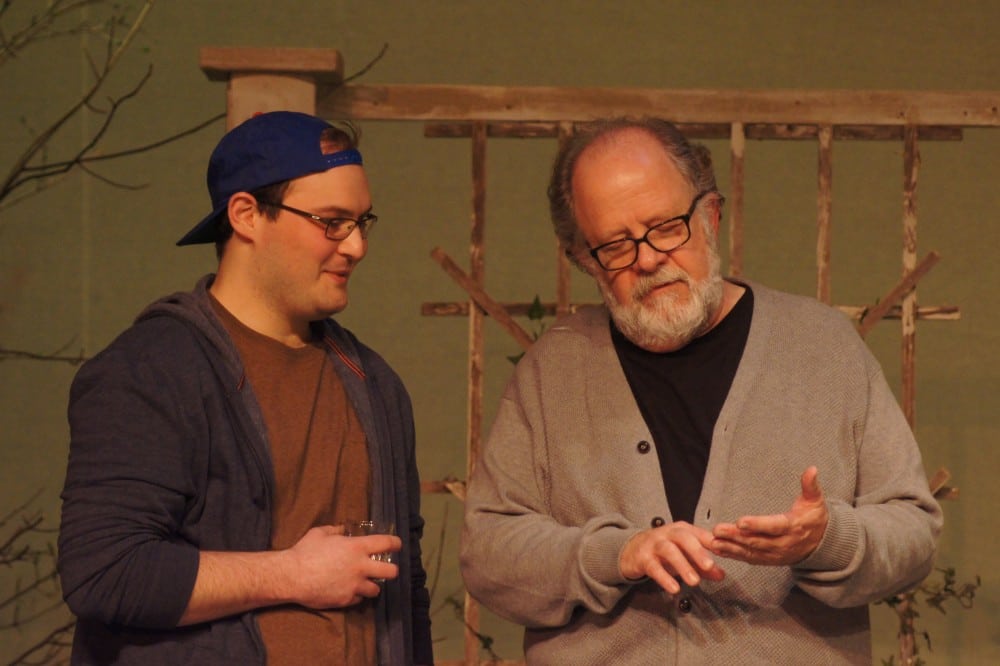 Two men stand together indoors. The older man with glasses and a gray sweater gestures as he talks, while the younger man in a blue cap and hoodie holds a glass and listens—capturing a scene from the award-winning show "Proof" at AAAA theatre.