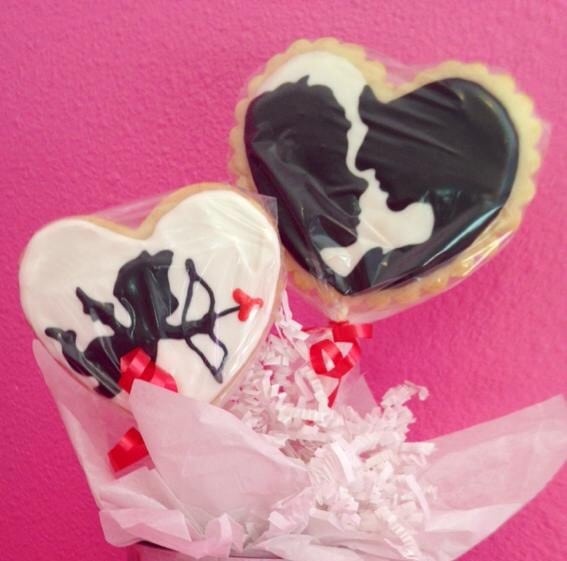 Two heart-shaped cookies on sticks, decorated with black and white icing—one featuring a couple’s silhouette, the other an abstract design with a red heart. Perfect for Valentine's Weekend in Alexandria, both are wrapped in plastic against a pink background.