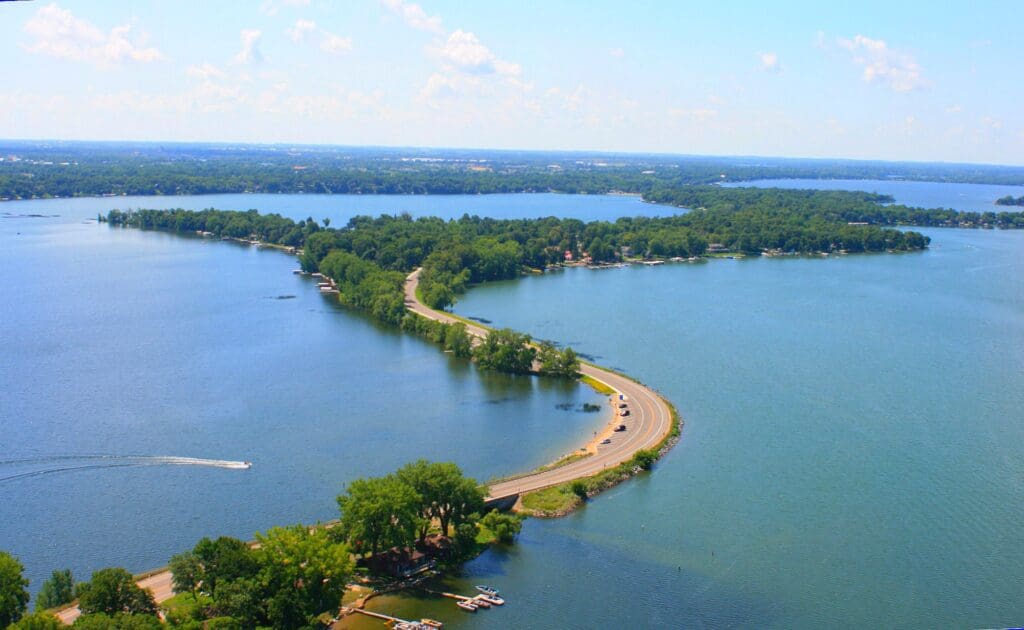 A winding road bordered by trees stretches across a narrow strip of land between two large blue lakes, with boats on the water and a bright, clear sky—one of the region’s must-see attractions.