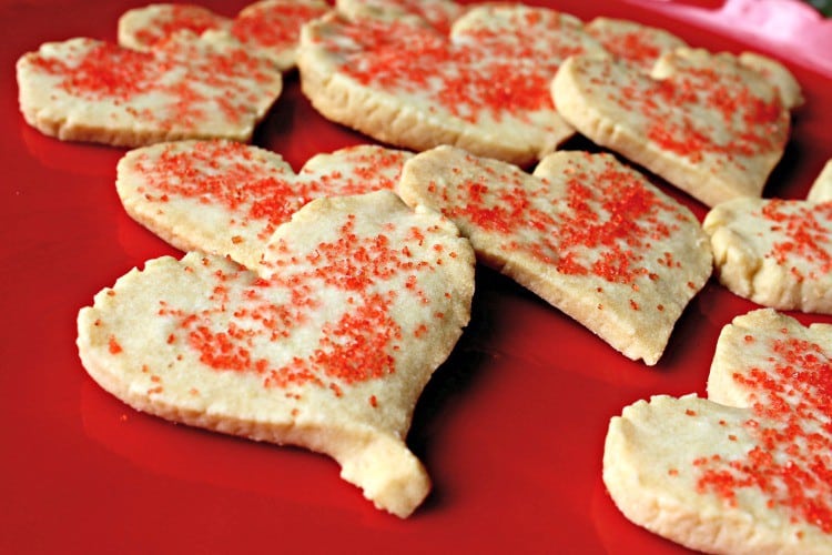 Heart-shaped sugar cookies topped with red sprinkles are arranged on a bright red surface, adding a touch of fun and romance perfect for Valentine’s Weekend.