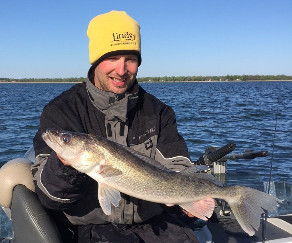 A smiling person in a yellow beanie and dark jacket holds a large early June Walleye while sitting in a boat on a lake with blue water and a tree-lined shore in the background.