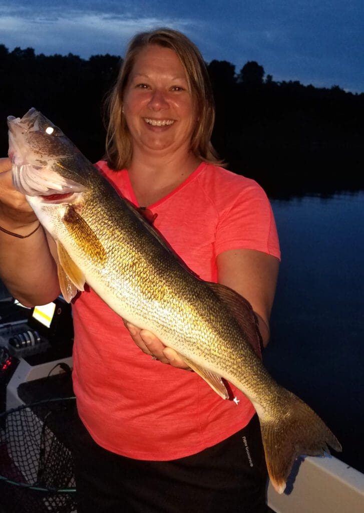 A smiling woman in a pink shirt proudly holds a large fish with both hands while standing on a boat at dusk, framed by dark water and trees in the background.