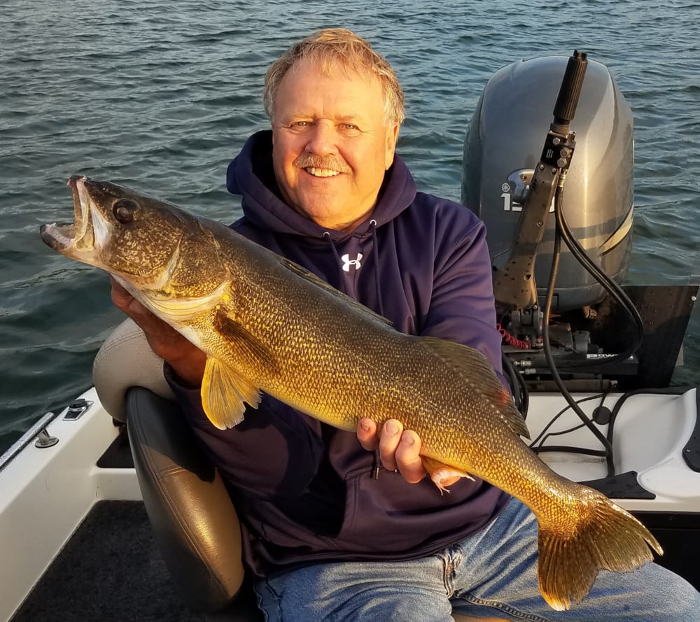 A smiling man in a navy hoodie sits on a boat, holding a large walleye fish with both hands—a perfect moment during summer fishing in Alexandria, MN. The background shows water and part of the boat's motor.