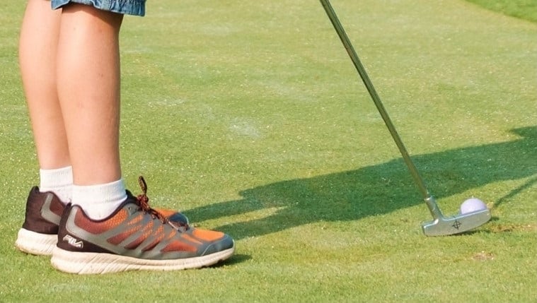 A person in sneakers and shorts stands on a green, preparing to hit a golf ball with a putter—a classic scene for anyone getting started in golf. Only their legs and the golf club are visible.