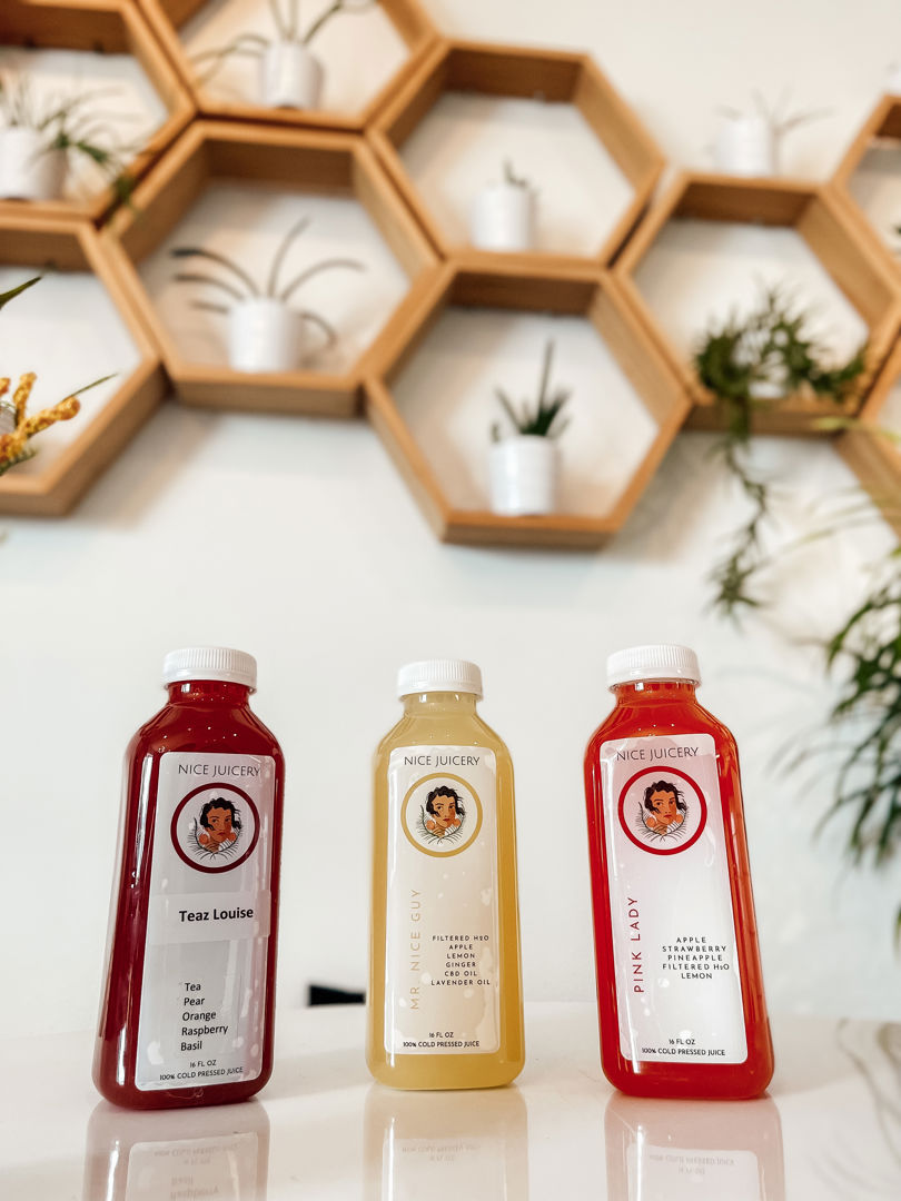 Three bottled drinks from Nice Juicery—labeled "Teaz Louise," "Mingle Gini," and "Pink Lady"—stand on a counter. Behind them, honeycomb-shaped wall shelves display plants and decor against a white wall.