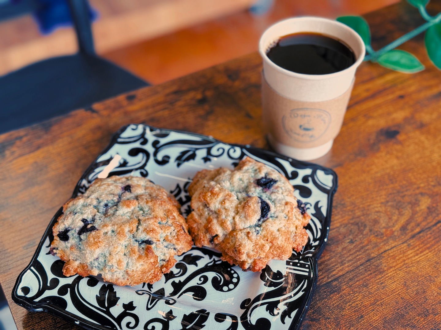 Two blueberry scones on a decorative black and white plate sit beside a cup of black coffee from Deja Bleu Coffee House, all arranged on a wooden table with a green plant partially visible.