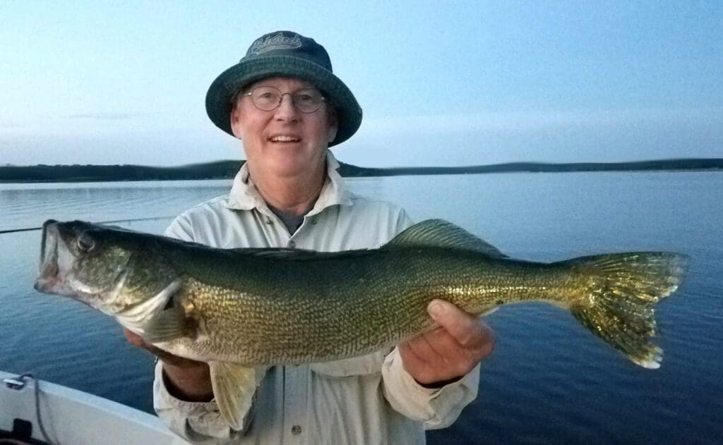 An older man wearing glasses and a bucket hat smiles while holding a freshly caught fish on a boat, with calm lake water and a distant shoreline in the background.