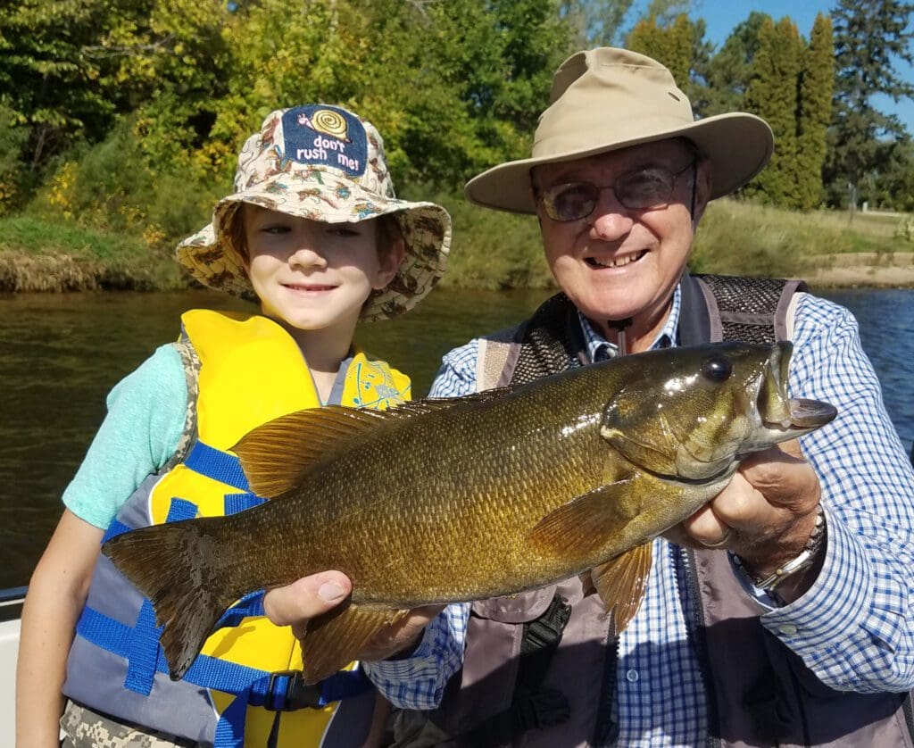 A smiling child in a yellow life jacket and an older man in a sun hat proudly hold a large fall fish together on a sunny day near a river, with green trees in the background.