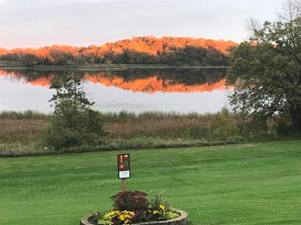 A grassy lawn at Osakis Country Club with a flower bed in the foreground overlooks a calm lake. Autumn trees with orange foliage are reflected in the water under an overcast sky, while a small sign stands near the flower bed.