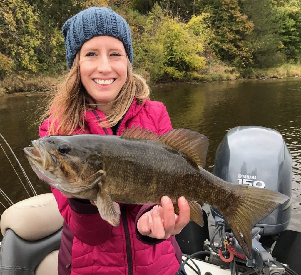 A smiling woman in a red jacket and blue knit hat proudly shows off her fishing catch on a boat, with a river and autumn foliage in the background.