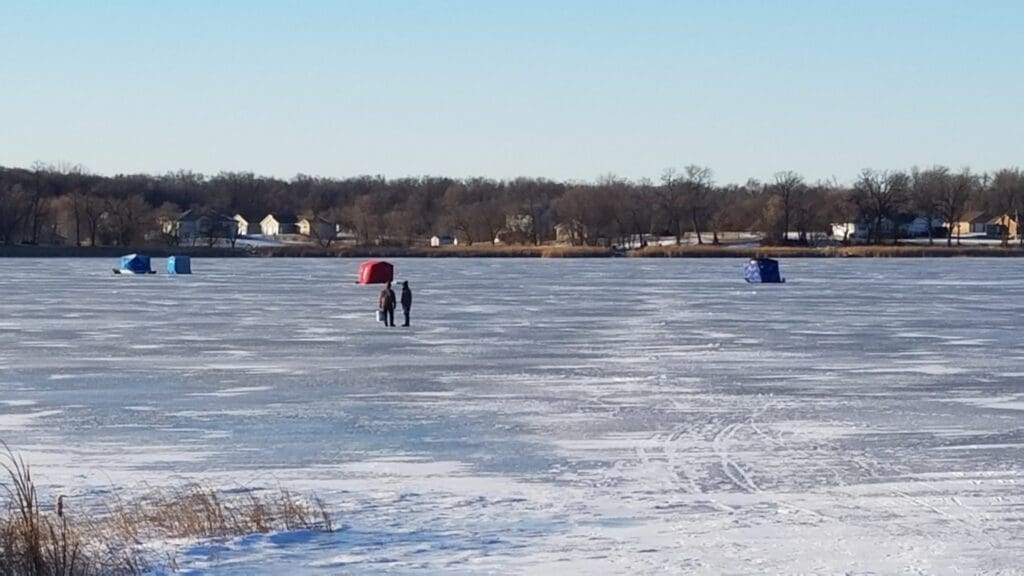 Two people walk on a frozen lake covered in snow and ice, heading toward colorful ice fishing shelters. Houses and trees line the far shore beneath a clear blue sky.