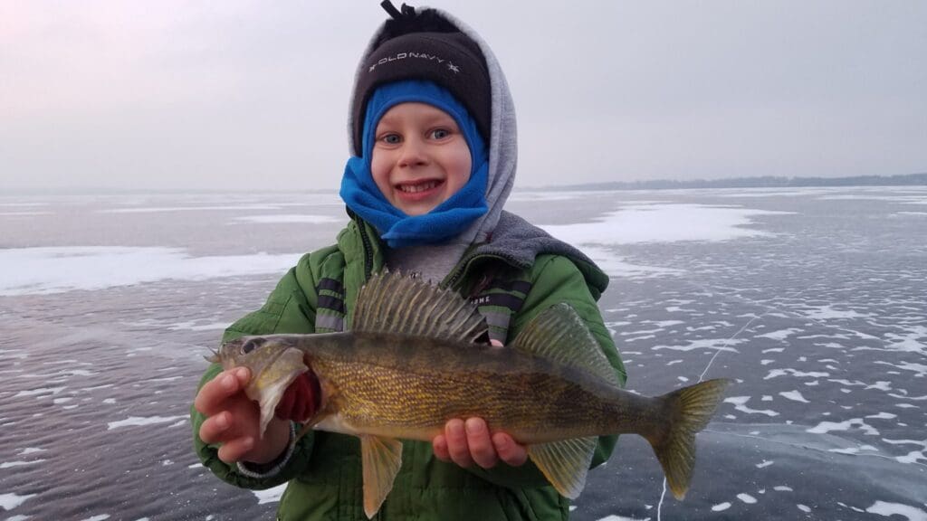 Smiling child in winter clothing holds a freshly caught fish while standing on a frozen, snow-dusted lake, proud of their December catch and ice report.