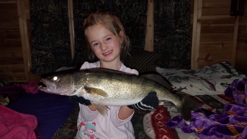 A young girl smiles as she holds a large fish with both hands, wearing gloves—perhaps after an exciting ice fishing adventure—indoors on a bed with patterned blankets.