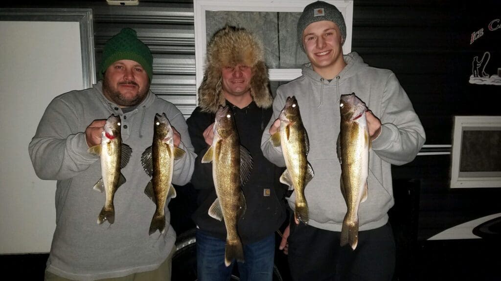 Three men dressed in winter clothing stand indoors, each proudly holding freshly caught fish from their fishing trip, smiling at the camera. The background includes a window and dark paneling.
