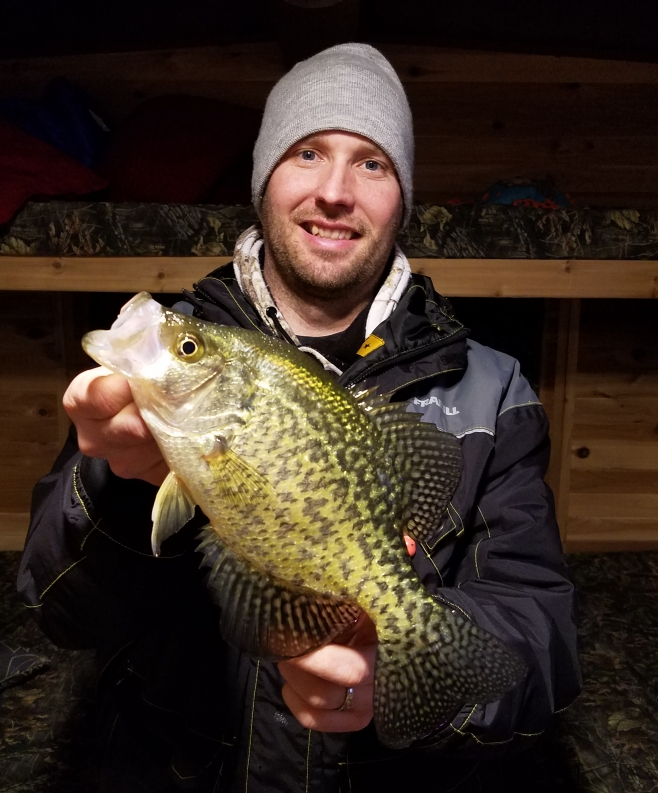 A smiling man wearing a gray beanie and a dark jacket proudly holds up his fishing catch—a large yellow and black speckled fish—indoors, with wooden bunk beds in the background.