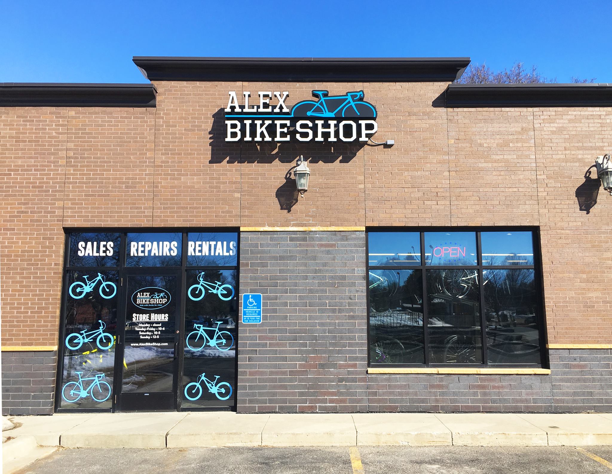 A brick storefront for Alex Bike Shop features a blue bicycle logo. Windows display bike graphics and the words “Sales, Repairs, Rentals.” An “Open” sign is lit inside, and there’s a handicap parking sign by the door.