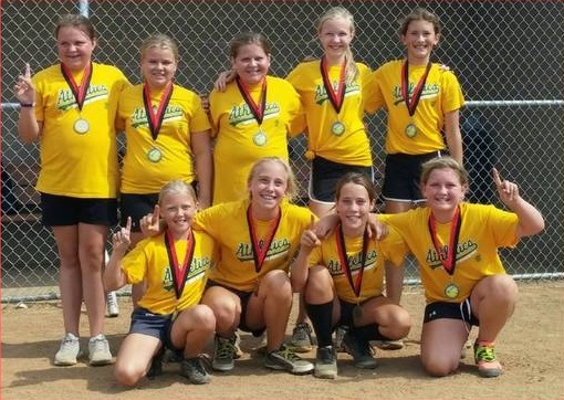 Nine girls in yellow "Athletics" shirts and medals pose on a dirt field, smiling and holding up one finger, proudly representing Alexandria Sports & Activity Organizations after winning a game or tournament.