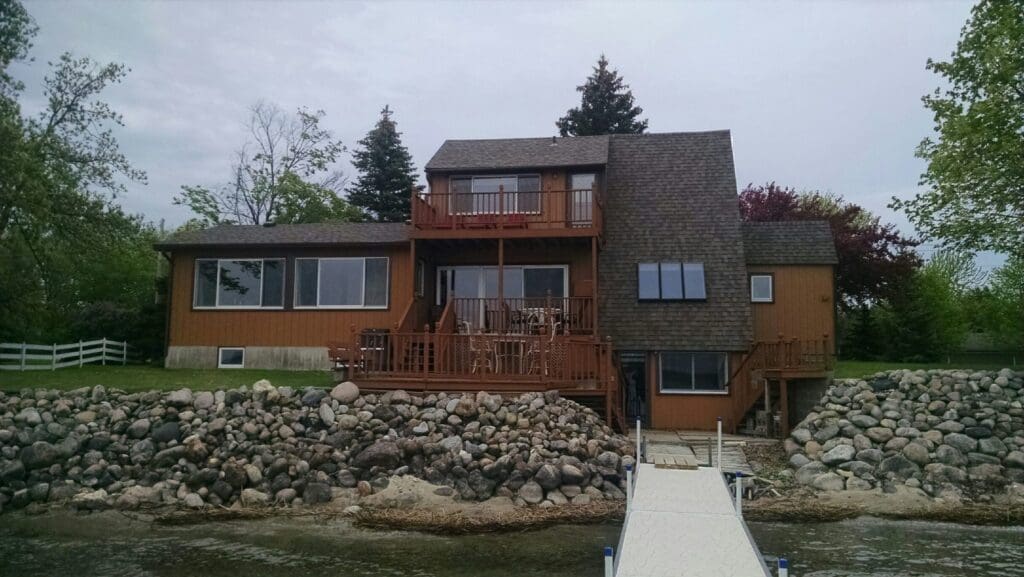 An A-Frame on Lake Darling with large windows and multiple balconies sits on a rocky shoreline. A white dock extends from the house over the water, surrounded by lush trees and greenery.