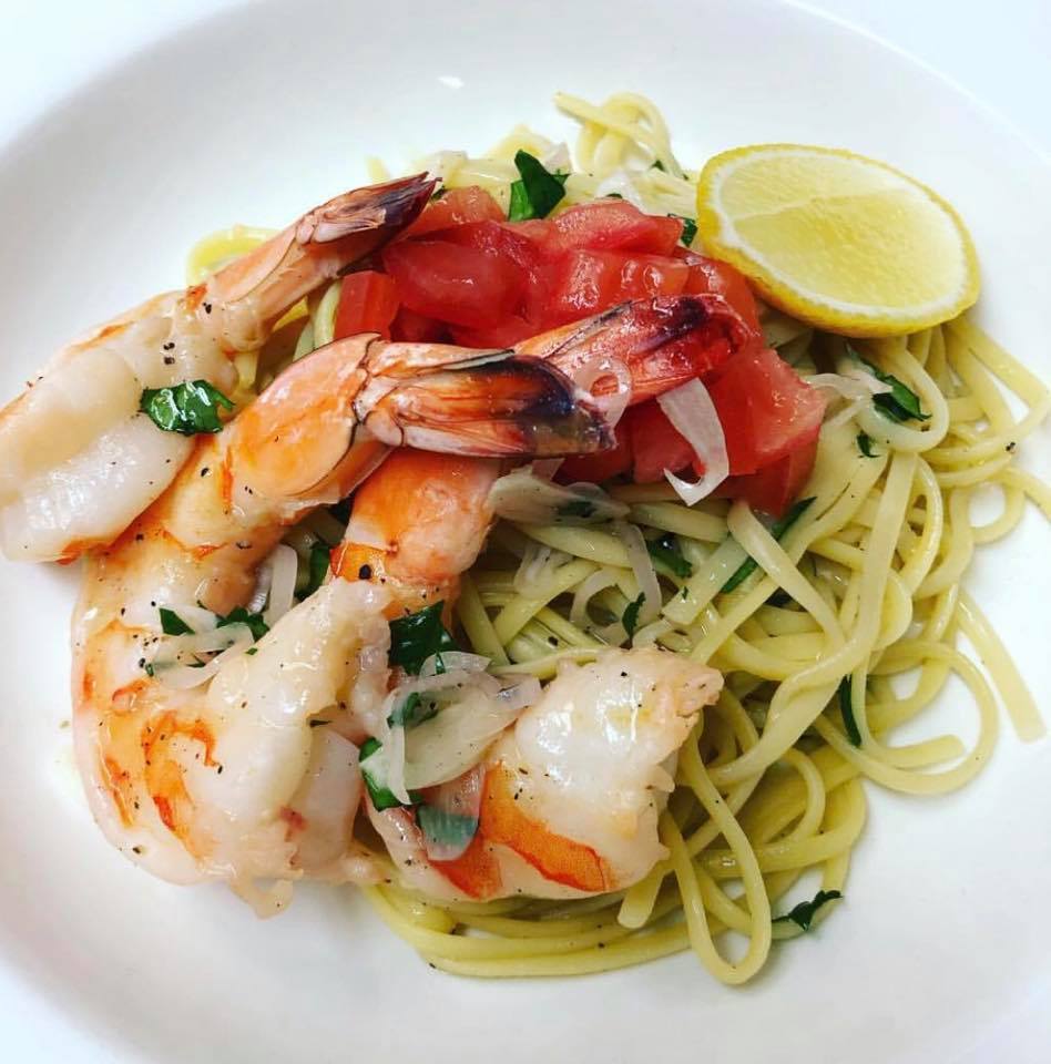 A dish of linguine pasta from Lure Lakebar, topped with grilled shrimp, diced tomatoes, chopped herbs, and onions, served with a lemon wedge on a white plate.