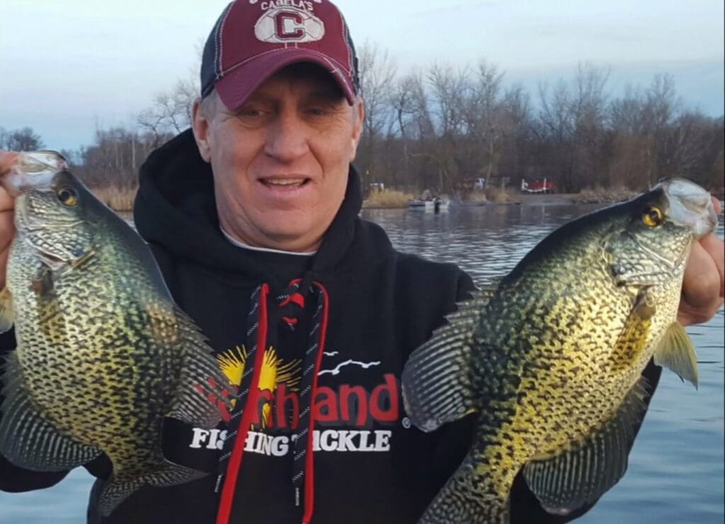 A man in a maroon cap and black hoodie holds up two large crappie fish, one in each hand, while fishing on a boat. Trees and water form the scenic background, reminiscent of an outing with Mike Frisch.