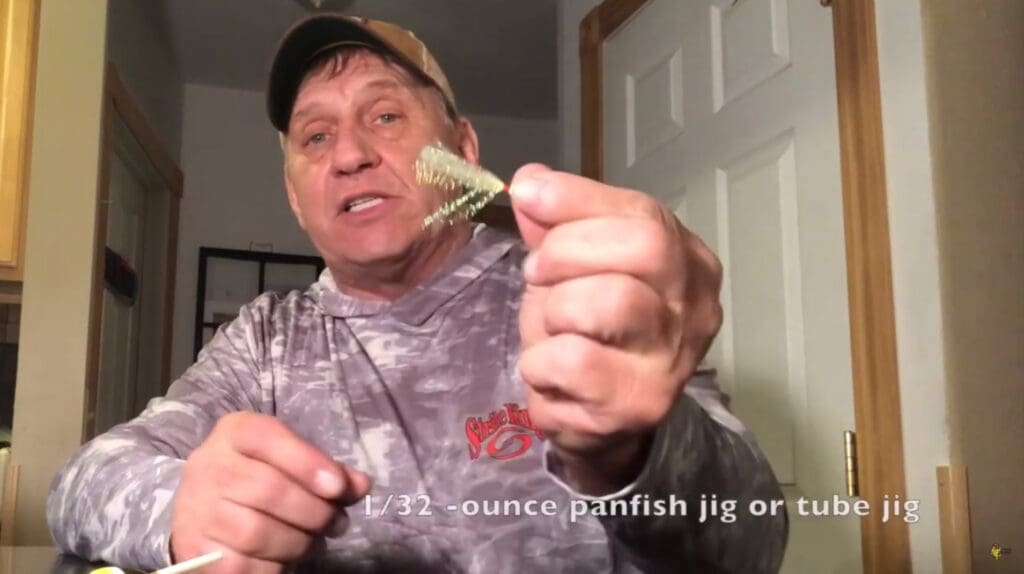 A man in a camo shirt and cap, possibly Mike Frisch, holds up a small fishing jig toward the camera. Text at the bottom reads: "1/32-ounce panfish jig or tube jig.