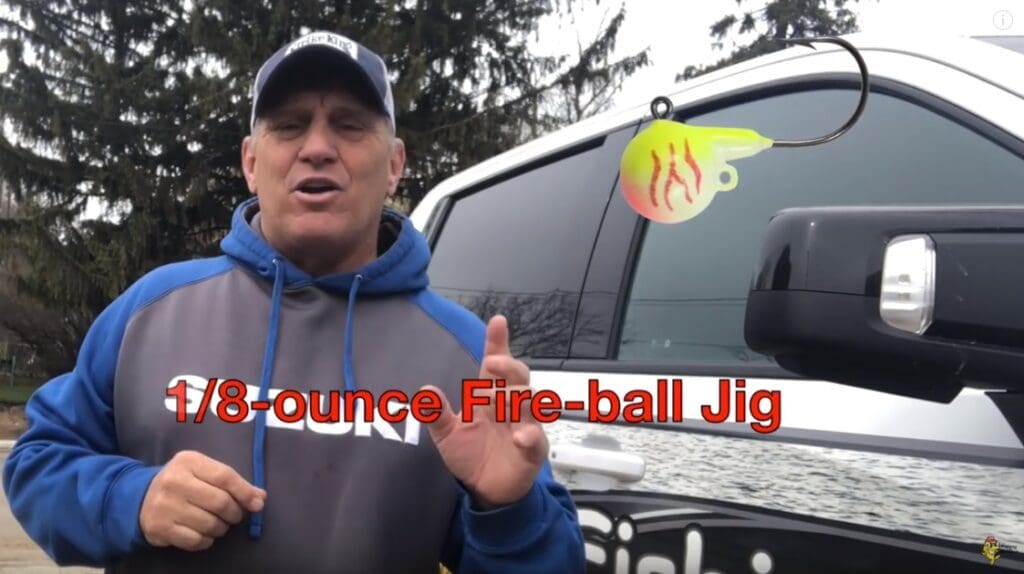 A man in a blue and gray hoodie stands next to a truck, holding up his finger. Overlaid text reads "1/8-ounce Fire-ball Jig" with an image of a yellow and red fishing jig above the text, featuring pro angler Mike Frisch.