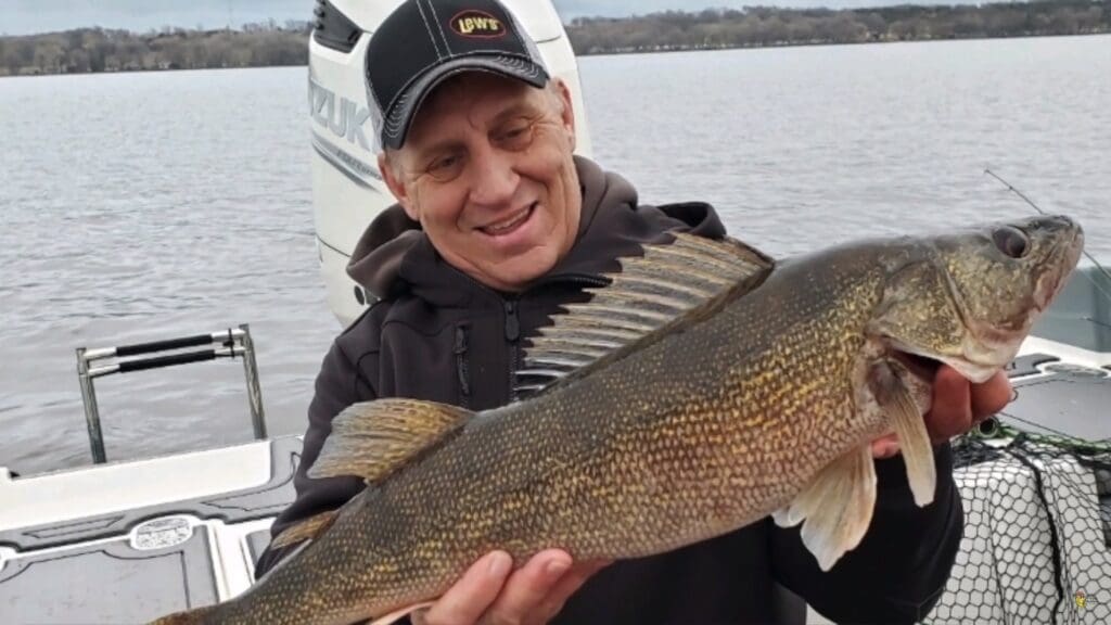 A smiling man in a black cap and hoodie holds a large walleye fish on a boat during fishing season, with a lake and shoreline visible in the background.