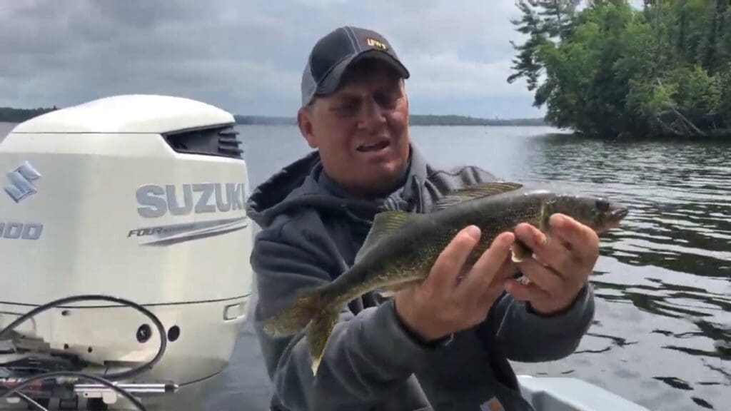 A man wearing a cap holds a fish with both hands on a boat near Alexandria. A Suzuki outboard motor and trees along the shore are visible in the background under a cloudy sky, making for a classic Mike Frisch fishing scene.