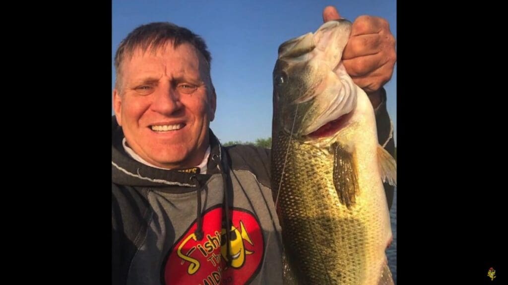 A smiling man in a gray hoodie with a red and yellow “Fishing” logo holds up a large fish under a clear blue sky. Mike Frisch proudly shows off his catch, capturing the perfect fishing moment.