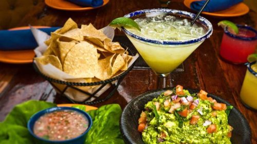A table at Rancho Viejo with a margarita, a basket of tortilla chips, a bowl of guacamole topped with diced tomato and onion, and a small dish of salsa, all on colorful plates.