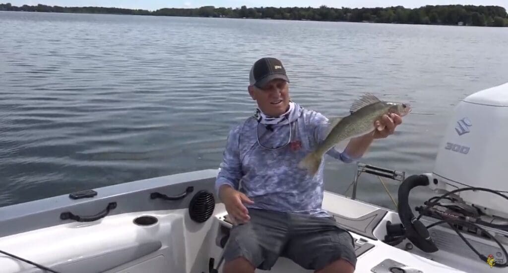 A man, possibly Mike Frisch, sits in a boat on a lake holding up his catch with a proud smile. He sports a cap, blue camouflage shirt, shorts, and a neck gaiter. Trees line the distant shore beneath a cloudy sky.