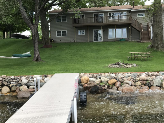A lakefront AA Cowdry Lake Home with a large grassy yard, hammock, picnic table, and fire pit. A wooden deck and stairs lead to the house, while a dock extends into the lake. Trees surround this peaceful property.