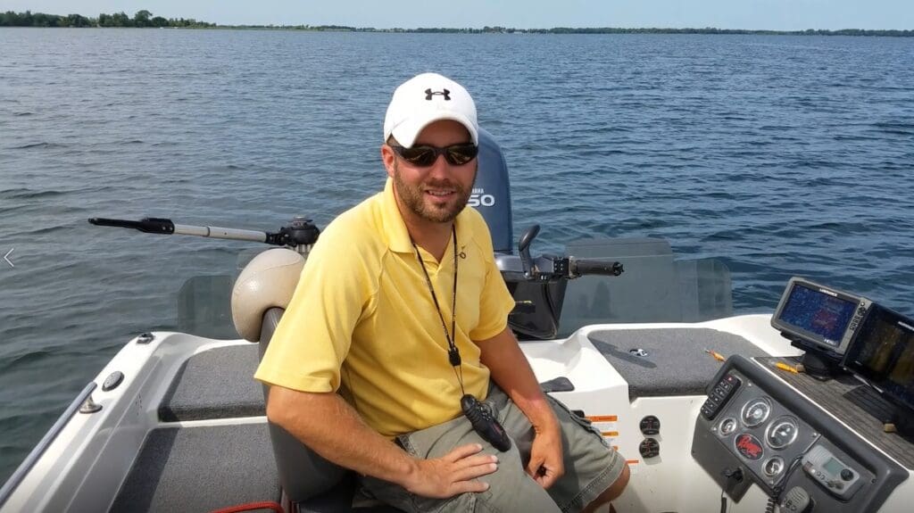 A man wearing a white cap, sunglasses, and a yellow shirt sits on a boat, smiling at the camera. Surrounded by fishing gear and boating equipment, he enjoys the day with a vast body of water and distant shoreline in the background.